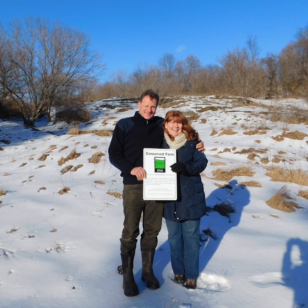 Couple standing in a snowy landscape, holding a "Conserved Farm" sign, celebrating their commitment to sustainable agriculture and land preservation.